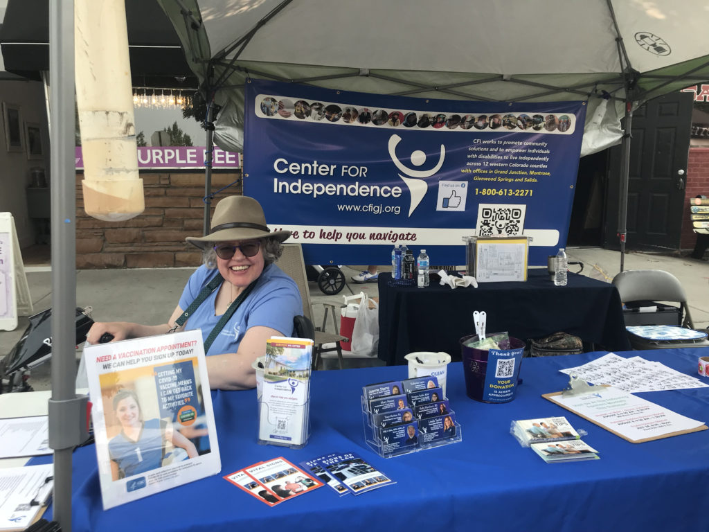 Woman with a hat on sits in a scooter chair sits behind a table at farmers market with brochures in front of her