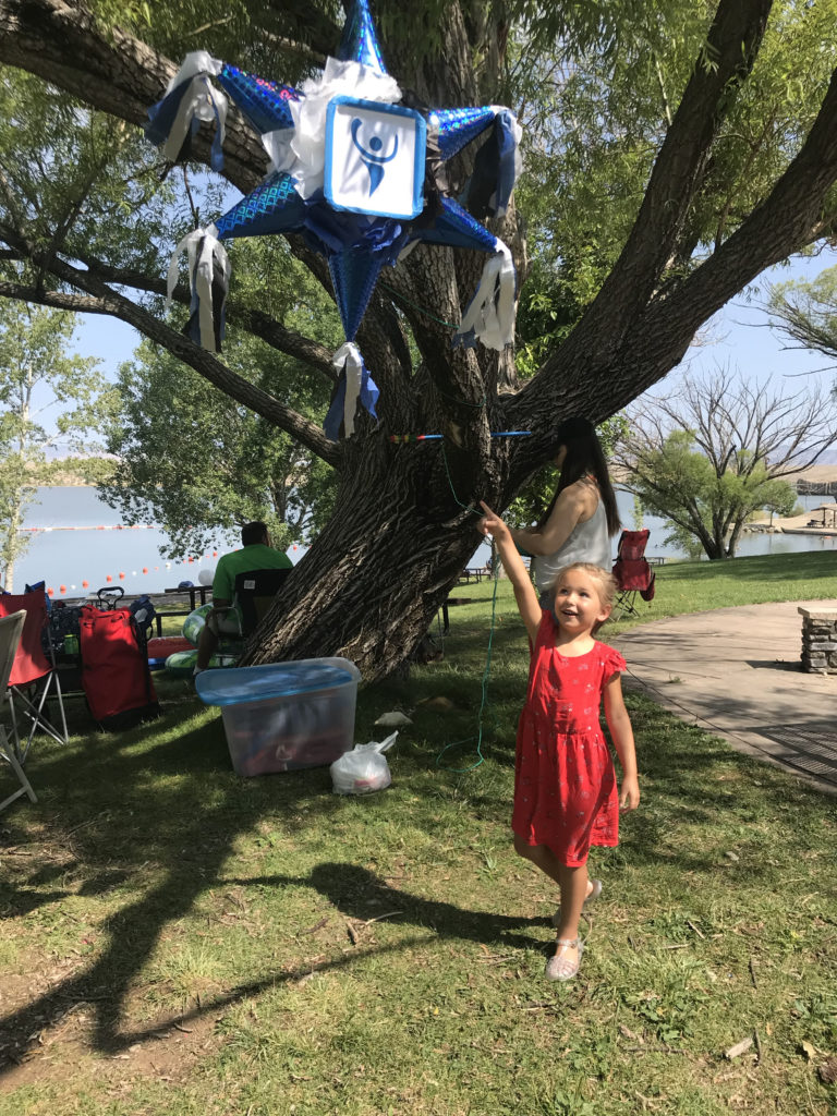Child in red dress points to a piñata hanging in tree above.