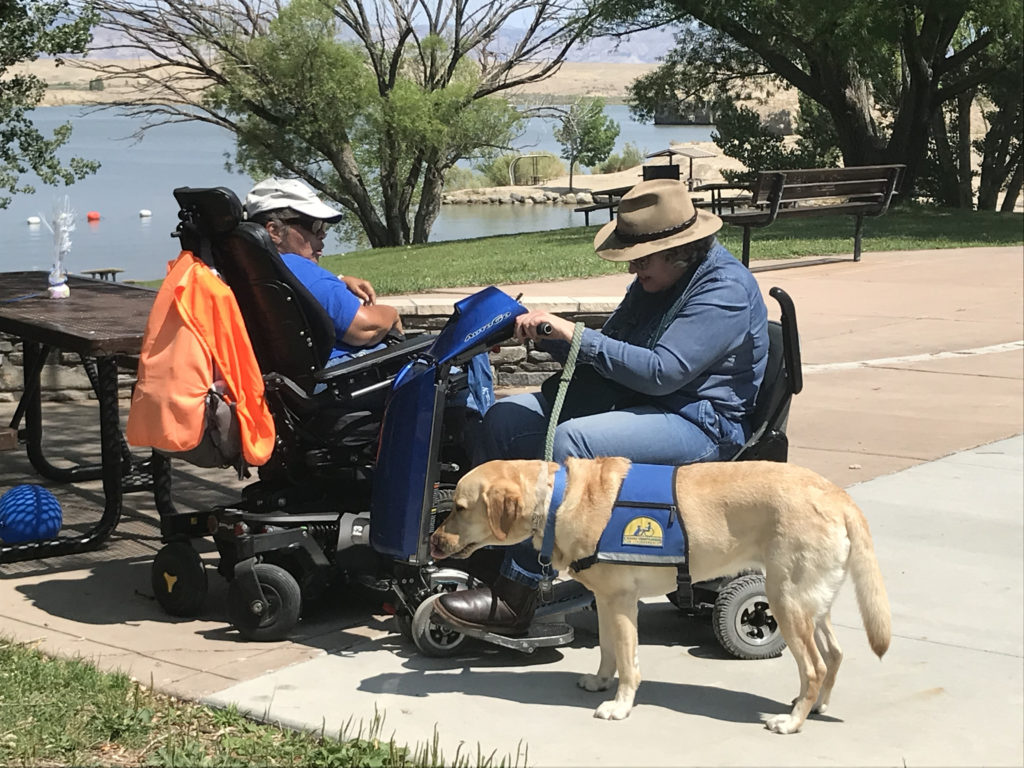 2 women in scooter wheelchairs talking with service dog.