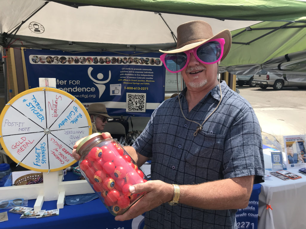 Man with hat and big pink sunglasses holds a jar of plastic eyeballs for "Guess How Many" challenge at event booth.